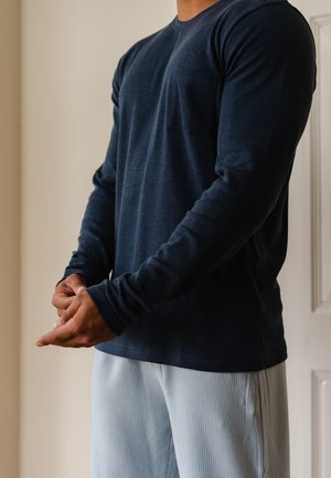 Navy long-sleeve t-shirt with a chest pocket, made of smooth fabric. Paired with light blue, pinstriped pants. Hands in a relaxed position.