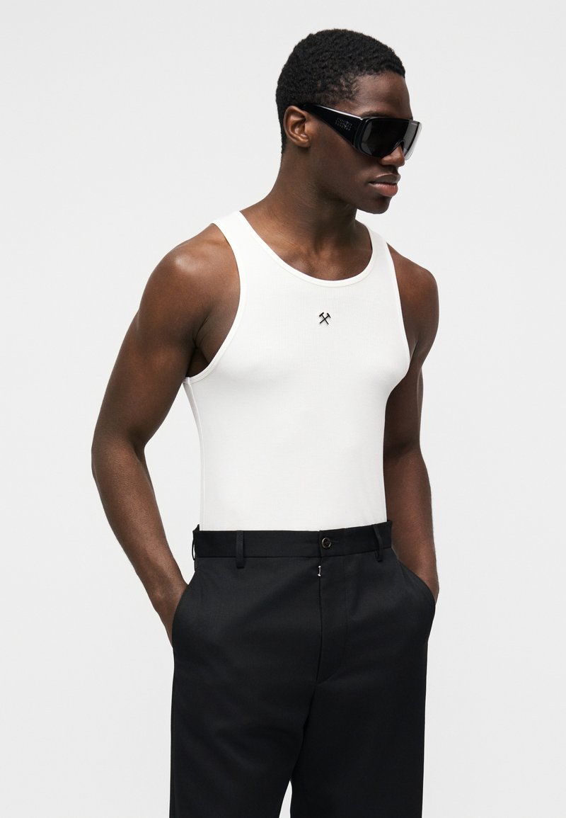 Débardeur blanc à côtes avec un col rond, associé à un pantalon noir ajusté. De grandes lunettes de soleil noires complètent le look.