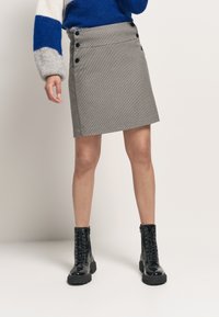 Szara spódnica w kratę z czarnymi guzikami w pasie, zestawiona z czarnymi botkami na sznurówki do kostek. Spódnica ma prosty krój i teksturowany materiał.