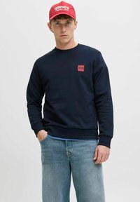 Marineblaues Sweatshirt mit einem kleinen roten Logopatch auf der Brust, kombiniert mit hellblauen Jeans und einer roten Baseballkappe.