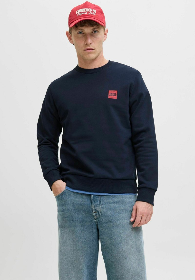 Marineblaues Sweatshirt mit einem kleinen roten Logopatch auf der Brust, kombiniert mit hellblauen Jeans und einer roten Baseballkappe.