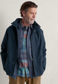 Chaqueta impermeable de color azul marino con capucha, que presenta un cierre de cremallera y botón a presión, llevada sobre una camisa de cuadros con botones. Textura suave y líneas limpias.