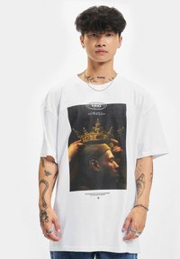 Hvid oversized t-shirt med en sort grafisk print af en kronet skikkelse, med tekst nedenfor og korte ærmer. Subtil struktur synlig.