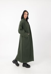 Langer, übergroßer grüner Mantel aus weichem Stoff, mit weiten Ärmeln, zwei Taschen und einer entspannten Passform. Kombiniert mit klobigen schwarzen Plattformschuhen.