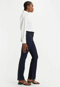 Witte button-up shirt, donkerblauwe slim fit jeans en zwarte sandalen met bandjes. De jeans hebben een lichte flare en een gladde denimtextuur.