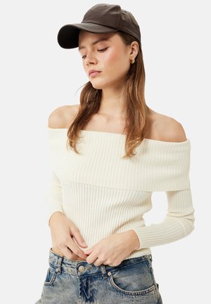 Ung kvinde med langt hår bærer en brun læderkasket, en cremefarvet ribstrikket trøje med bare skuldre og blå jeans, ser ned med hænderne nær taljen.