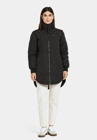 Svart vadderad lång kappa med hög krage, dragkedja och ribbade muddar, kombinerad med krämfärgade byxor och vita sneakers.