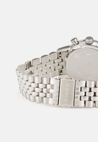 Montre en acier inoxydable argenté avec un bracelet à maillons et un boîtier rond. Présente un dos texturé et une marque gravée sur la fermeture.
