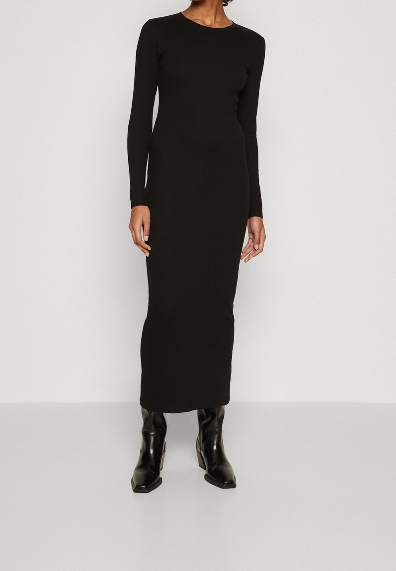 Svart långärmad klänning med en figursydd silhuett och rund halsringning, tillverkad av ett mjukt tyg. Matchas med höga svarta klackstövlar.