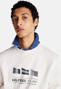Sudadera blanca con un gráfico de tres banderas y el texto "HILFIGER Est. 1985." Capas con un cuello de mezclilla azul y una camisa abotonada debajo.