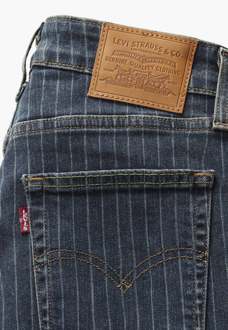 Jean en denim foncé avec des rayures verticales, une étiquette en cuir marron avec un logo embossé et une étiquette rouge sur la poche arrière.