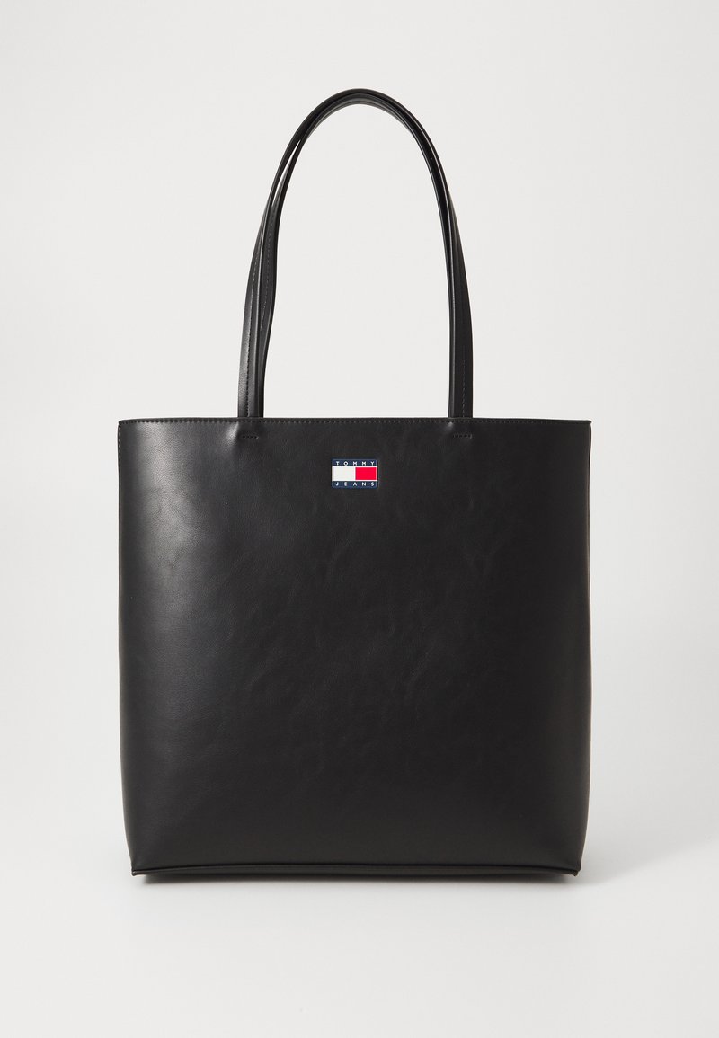 Sac fourre-tout en cuir noir avec deux longues poignées, au design minimaliste et un petit logo cousu à l'avant. Texture lisse, forme rectangulaire.