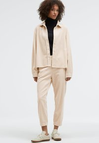 Beige strukturierte Langarmhemd mit Knöpfen, kombiniert mit passenden Hosen und hellen Sneakers. Schwarzes Rollkragenoberteil ist sichtbar darunter.