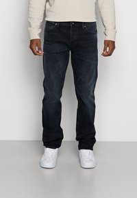 Dunkelblaue Jeans mit schmaler Passform, fünf Taschen und dezenten ausgewaschenen Akzenten. Kombiniert mit weißen Sneakers vor einem einfarbigen Hintergrund.