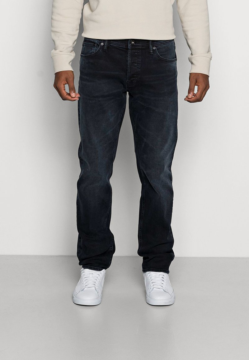 Dunkelblaue Jeans mit schmaler Passform, fünf Taschen und dezenten ausgewaschenen Akzenten. Kombiniert mit weißen Sneakers vor einem einfarbigen Hintergrund.