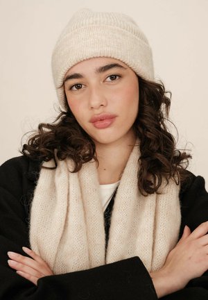 Un bonnet en tricot beige clair avec une texture en côtes, associé à une écharpe crémeuse douce et pelucheuse. Le mannequin porte un manteau noir.