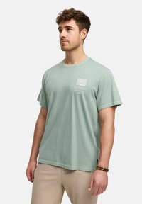 T-shirt en coton vert menthe à manches courtes, col rond, avec un imprimé graphique sur la poitrine gauche, associé à un pantalon beige plissé.