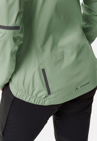 Giacca impermeabile verde chiaro con vita elastica, accenti neri e un logo. Progettata per l'uso all'aperto, mostra un aspetto funzionale e slanciato.