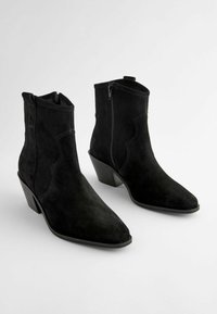 Botas de tobillo de ante negro con punteras afiladas, cremalleras laterales y tacones apilados. Presenta detalles de costura para un acabado adicional.