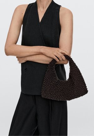 Kobieta w czarnej kamizelce bez rękawów i spodniach trzyma czarną, fakturowaną torebkę na koraliki z zaokrąglonym uchwytem na jednolitym tle.