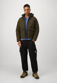 Zöld steppelt dzseki kapucnival, két mellzsebbel és elülső cipzárral. Kék ing felett viselve, fekete cargo nadrággal és barna cipővel.