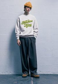 Felpa bianca con la scritta "Hawkins Tigers" in corsivo verde. Pantaloni grigi a pieghe e scarpe marroni con lacci blu. Berretto di lana arancione.