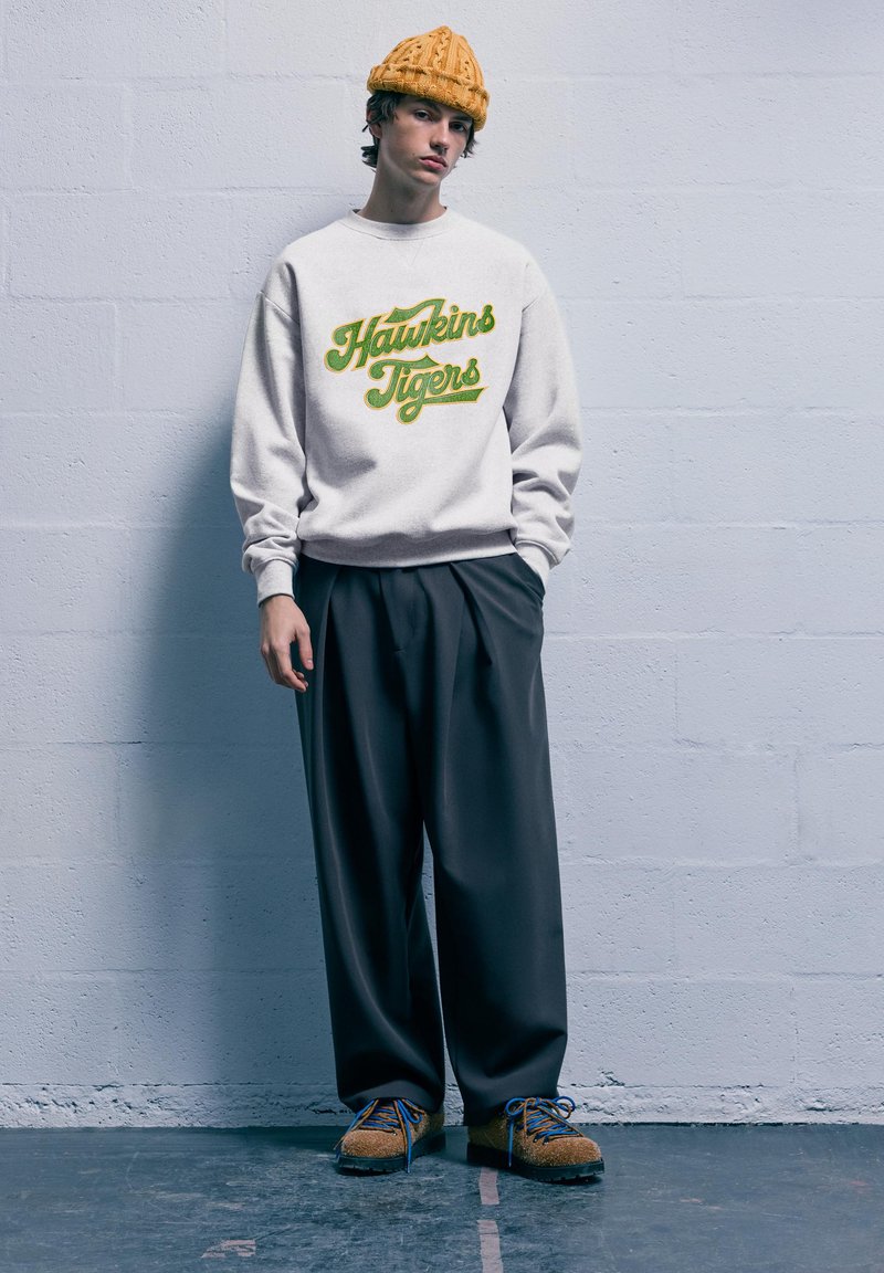 Felpa bianca con la scritta "Hawkins Tigers" in corsivo verde. Pantaloni grigi a pieghe e scarpe marroni con lacci blu. Berretto di lana arancione.