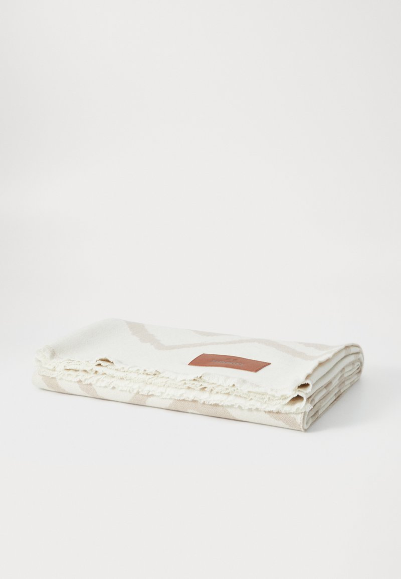 Krém színű, finom bézs mintákkal ellátott szőtt takaró, fröccsöntött szélekkel és egy téglalap alakú bőr címkével, amelyen a márkaneve található.