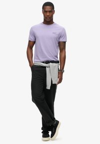 Maglietta corta viola, pantaloni neri, maglione grigio annodato in vita, sneakers nere con suola crema, persona in piedi con atteggiamento sicuro.