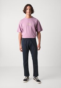 Světle fialové bavlněné tričko s logem „Rip Curl“, spárované s černými slim-fit kalhotami a černobílými nízkými teniskami.