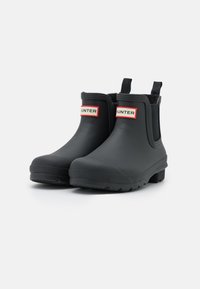 Bottes de cheville en caoutchouc noir avec une finition mate, panneaux latéraux élastiques et marquage "HUNTER" en rouge à l'avant. Talon bas et semelle texturée.