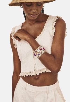 Top corto blanco con borde de encaje, adornado con botones. Brazalete dorado con detalles florales en colores. Combinado con pantalones a juego.