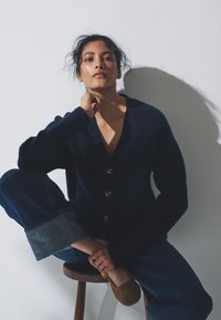 Granatowy sweter rozpinany z dekoltem w serek i pięcioma guzikami, w połączeniu z ciemnymi dżinsami, które mają podwinięte mankiety, oraz brązowymi butami wsuwanymi.