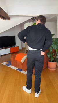 Mann trägt schwarzen Pullover und Hose und richtet seine Kleidung im Wohnzimmer mit orangefarbenem Sofa und großer Topfpflanze.