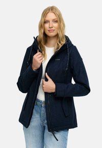 Femme portant une veste à capuche bleu marine sur une chemise blanche et un jean bleu clair, tenant le col et la fermeture éclair de la veste avec les deux mains.