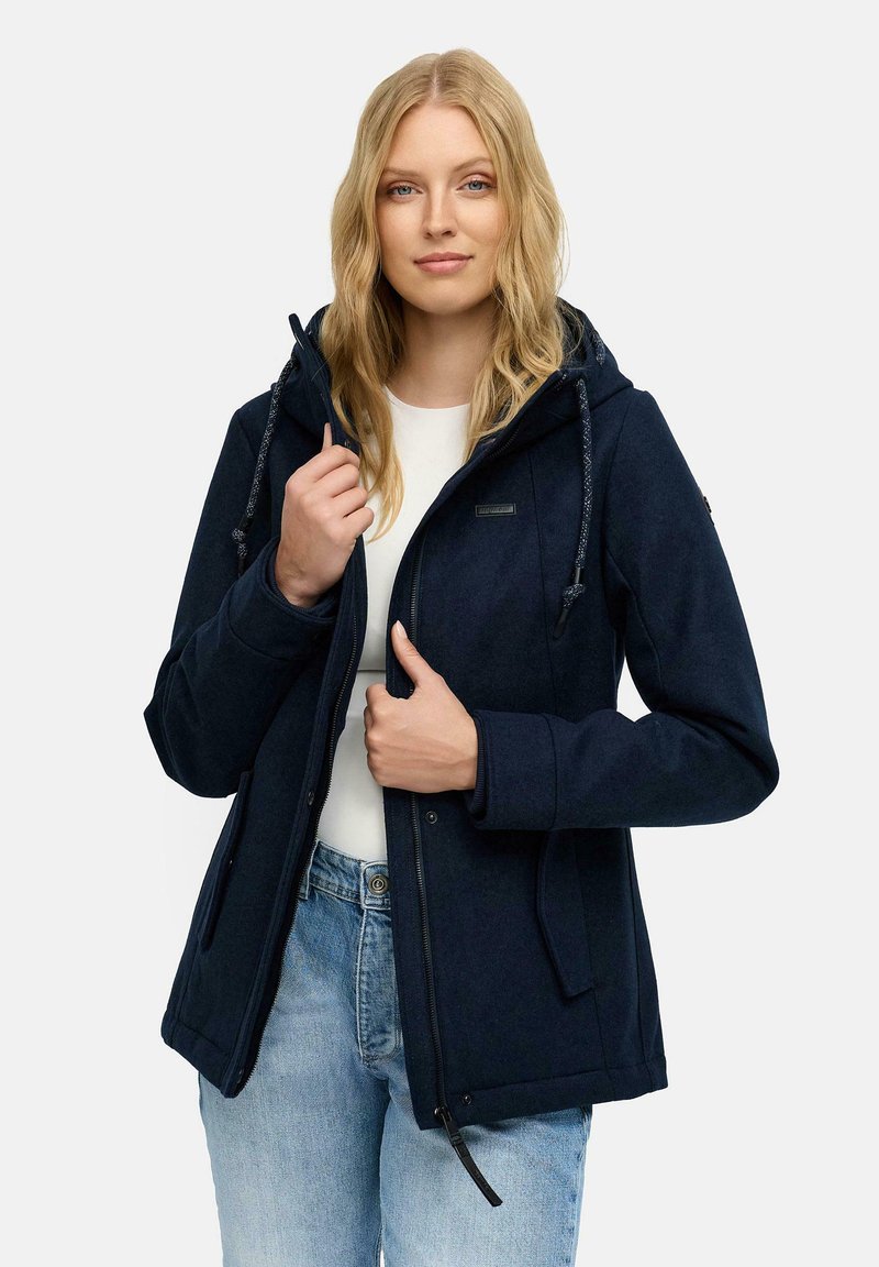 Femme portant une veste à capuche bleu marine sur une chemise blanche et un jean bleu clair, tenant le col et la fermeture éclair de la veste avec les deux mains.