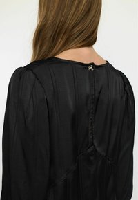 Blusa in satin nero con pieghe verticali, scollatura rotonda e chiusura con bottone sul retro. Presenta maniche a sbuffo e texture liscia.