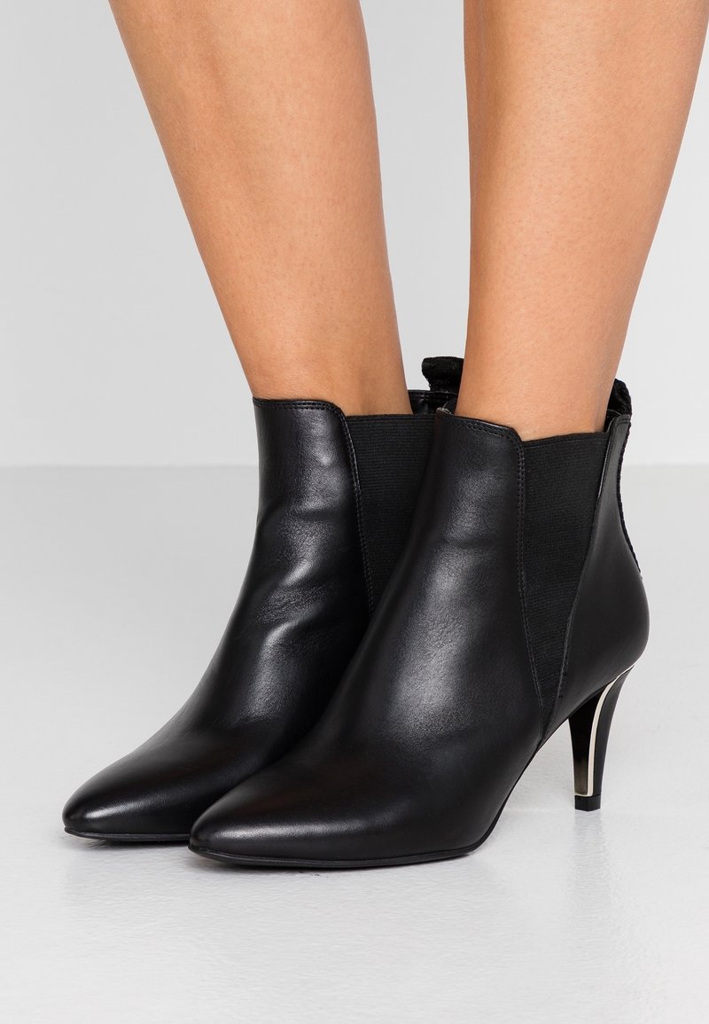 Svarta läderankelstövlar med spetsig tå, elastiska sidopaneler och metallisk stilettklack på en slät vit yta.