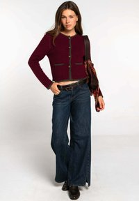 Cárdigan corto burdeos con ribete verde y dos bolsillos frontales, combinado con jeans de pierna ancha azules. Un cinturón en tono madera y zapatos oscuros completan el conjunto.