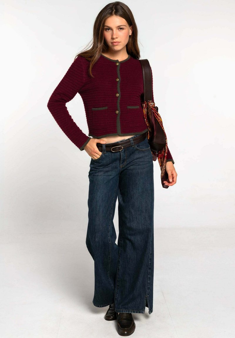 Cárdigan corto burdeos con ribete verde y dos bolsillos frontales, combinado con jeans de pierna ancha azules. Un cinturón en tono madera y zapatos oscuros completan el conjunto.