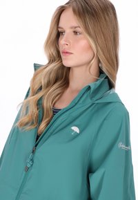 A zöldeskék vízálló dzseki cipzáras elülső résszel, állítható kapucnival és fehér esernyő logóval a mellrészen. Simaságú anyag és kényelmes, laza szabás.
