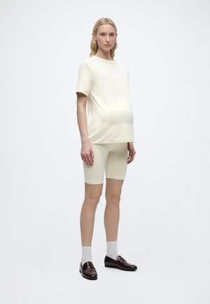 Schwangere Frau trägt cremefarbenes kurzärmeliges Oberteil und passende Biker-Shorts mit weißen Socken und dunklen Loafers, steht vor einem schlichten weißen Hintergrund.