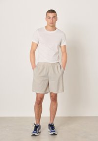 Młody mężczyzna w białej koszulce, beżowych szortach z kieszeniami oraz niebiesko-czarnych butach do biegania, stojący na jednolitym tle.