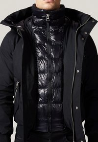 Czarna kurtka zapinana na zamek błyskawiczny z elegancką, połyskującą, pikowaną warstwą wewnętrzną. Projekt z kołnierzykiem z błyszczącymi akcentami, teksturowanymi panelami i bocznymi kieszeniami.
