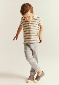 Gestreepte polo in beige en groen, gecombineerd met lichtgrijze joggers. De schoenen zijn grijs met een crème zool. Het kind staat met één voet omhoog.