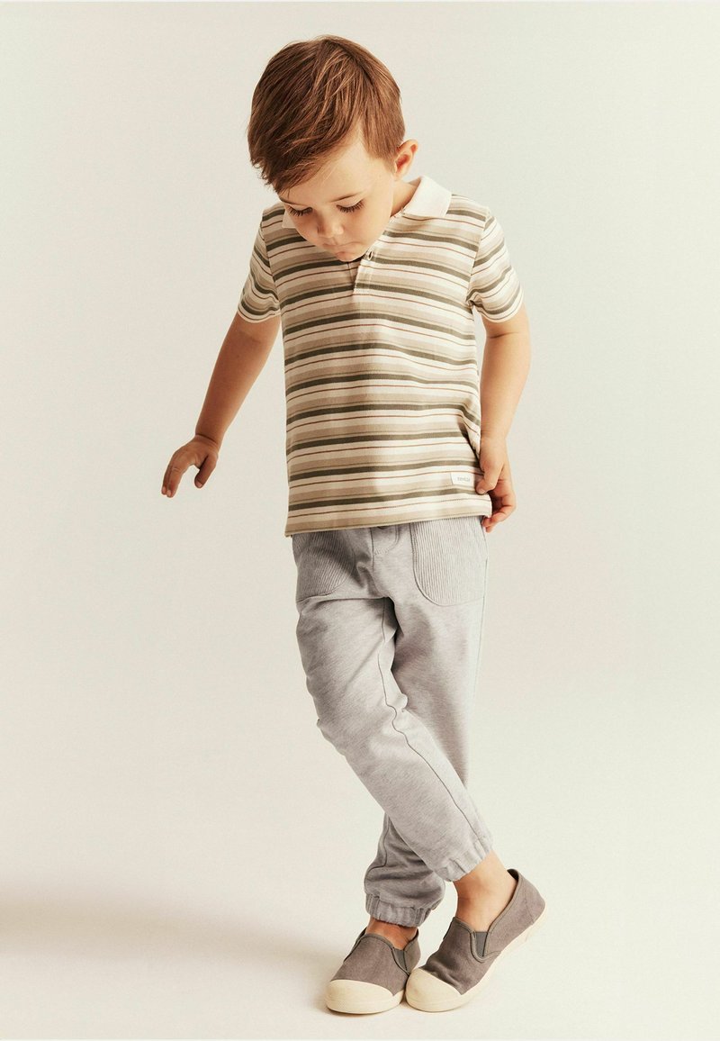 Gestreepte polo in beige en groen, gecombineerd met lichtgrijze joggers. De schoenen zijn grijs met een crème zool. Het kind staat met één voet omhoog.