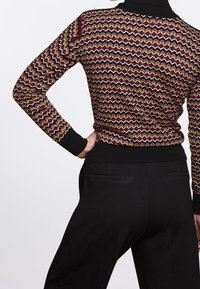 Pull en tricot présentant un motif en zigzag en bordeaux, jaune et noir. Col montant et ourlet côtelé, associé à un pantalon noir sur mesure.