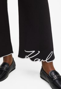 Melni bikses ar viļņotu apakšmali, kurai ir balti šķērsšuvumi un vārds "Karl" izsistā rakstā. Apvienotas ar melnu ādas plakanām kurpēm.