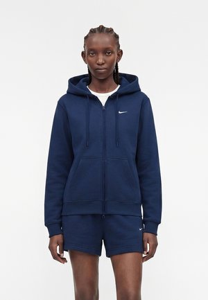 Femme aux cheveux tressés portant un sweat à capuche bleu marine zippé et un short assorti avec des logos Nike blancs, debout devant un fond blanc uni.