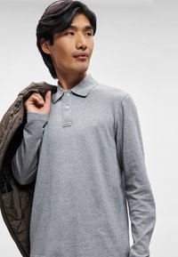 Polo shirt a maniche lunghe in grigio chiaro con colletto piatto, caratterizzato da tre bottoni e un piccolo logo sul petto. Tessuto dalla consistenza liscia e morbida.